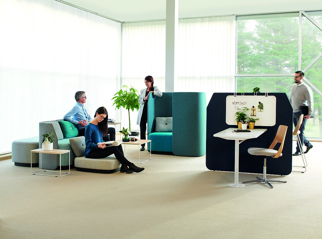 Ein modern eingerichteter, offener Büroraum. Es sind mehrere Polstermöbel und textile Trennwände zu sehen. Auf dem Boden liegt Teppich. 